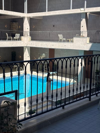 This media file depicts an outdoor swimming pool area, likely at a hotel or resort, based on the architectural style and the presence of lounge chairs. The pool is filled with clear blue water, and a shadow of a figure can be seen in the pool, suggesting someone is swimming or has recently been in the water. A person in white clothing is lying on a lounge chair beside the pool, partially visible.
The scene is viewed from above, looking down onto the pool and surrounding balconies or walkways. A black wrought-iron railing runs along the foreground, obscuring some of the view but also adding to the aesthetic of the space. The architecture features light-colored stone or tile cladding on the walls, with clean, modern lines.
Sunlight is casting strong shadows, indicating it is likely daytime, possibly mid-morning or afternoon, given the angle of the shadows. The weather appears to be clear and sunny. The location is specified as Ayn al Basha, Jordan. No visible text is present in the image. The overall impression is one of a relaxing and pleasant leisure environment.
