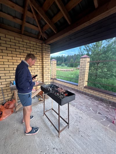 A man stands on a patio next to a barbecue grill, holding a mobile phone and a small white object. He is wearing a dark blue jacket, blue shorts, and dark blue Crocs. The barbecue grill contains burning coals and meat on a rack. The patio is paved with stone and concrete. Behind the patio is a brick wall and a wooden roof structure. In the background, there is a lawn, trees, and a forest. The scene appears to be outdoors, possibly in a backyard or a rural area. The sky is cloudy.