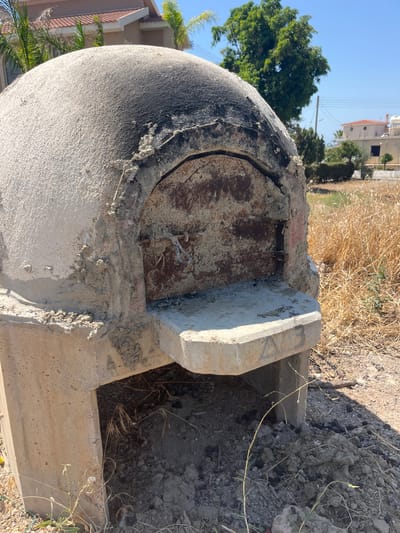 A rustic, dome-shaped oven made of concrete and plaster sits in an outdoor environment. The oven is supported by a concrete base with an opening underneath. The dome has a weathered appearance, with areas of soot near the top and around the opening. A metal door, rusted and grimy, covers the oven's mouth. The ground around the oven is covered in dry grass, dirt, and small stones. In the background, there are buildings with terracotta roofs and lush green trees under a clear blue sky. The location is identified as Moni Community, Cyprus, suggesting a possibly rural or semi-rural setting with residential structures nearby.