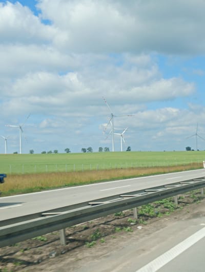 The image depicts a roadside view with a highway in the foreground and a field with wind turbines in the background. The highway is paved and has a metal guardrail. The field is green with tall grass and scattered trees. Several wind turbines are visible in the distance, with their blades stationary. The sky is partly cloudy with blue patches. The scene appears to be in a rural area. The location is identified as Kostomłoty, Poland.