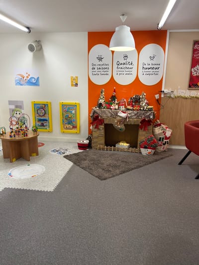 The image shows a brightly colored children's play area. A faux fireplace is decorated with Christmas decorations, including stockings and wrapped gifts. The walls are adorned with colorful graphics and signage in French, promoting fresh, seasonal food. Toys such as bead mazes and wall-mounted activity boards suggest a focus on early childhood education and entertainment. The flooring is a mix of gray carpet and interlocking foam tiles, offering a comfortable and safe environment for kids to play.