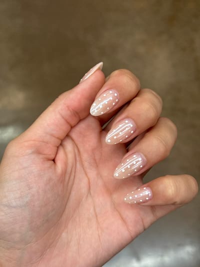 A human left hand is depicted in a close-up, dorsal view, presenting manicured fingernails against a uniformly blurred, light brown background. The nails are elongated, with an almond shape, and feature a translucent light beige or nude base coat. The index, middle, and pinky fingernails are adorned with a pattern of small, evenly spaced white polka dots. The ring finger nail displays horizontal white stripes. The thumb is partially visible at the lower right. No other subjects or objects are present. The hand is still, with fingers slightly splayed. The environment lacks specific identifying features, and no location within Shingle Springs, United States, can be ascertained from the visual data.