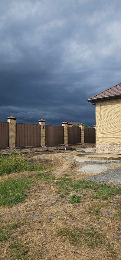 This media file depicts a view of a residential building with a metal roof and brick facade, alongside a fence and a patch of dry grass with some green vegetation. The building appears to be a home or a similar structure, and the fence suggests a property boundary. The scene is set outdoors, with a significant portion of the frame occupied by a dark, overcast sky, indicating potentially stormy weather. The presence of dry grass suggests a warm or dry climate, possibly during a summer or late autumn season. The location is identified as Овощной, Russia. There is no visible text in the image. The image is oriented sideways.