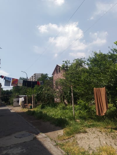 In the city of Nor Hachin, Armenia, this image captures a sunny day with a clear blue sky dotted with white clouds. The scene depicts an outdoor laundry area. Several clotheslines are strung horizontally, one above another, holding a variety of laundered items, including colorful towels, dark pants, and white shirts. A large, orange-brown curtain hangs from a clothesline on the right side of the frame, secured by clothespins. The background features lush green trees and a pink brick building with a visible roofline and windows. A multi-story building under construction is visible further in the distance. The foreground shows a paved path or road alongside a patch of unkempt grass and dirt.