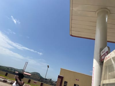 A person with dark dreadlocks, wearing a white tank top, stands at a gas station in Pasere, Nigeria, wiping their forehead. The sky is clear blue with scattered white clouds. A concrete pillar supports the roof of the gas station canopy, which has a beige ceiling and a red trim. A sign on the pillar reads "Shafa PMS 4". In the background, a light yellow building with a red trim has the word "MASJID" written on its exterior. Other buildings and structures, including some with red roofs, are visible further in the distance, along with rolling green hills. The ground appears to be paved with some areas of grass and concrete barriers. A street light stands in the middle ground.