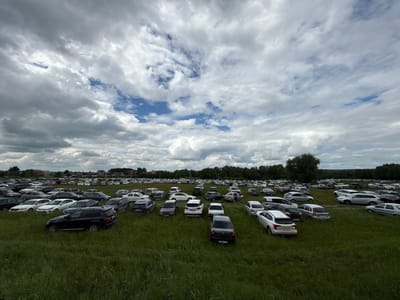 This is a wide-angle, eye-level photograph of a large outdoor parking lot filled with numerous cars parked on a grassy field under a cloudy sky. The parking lot appears to be located in a rural or semi-rural area, with trees and some buildings visible in the background. The cars are of various makes and models, and they are parked in rows, taking up a significant portion of the frame. The grass is tall and green, indicating a summer or late spring season. The sky is dramatic with a mix of dark and light grey clouds, with patches of blue sky visible through the gaps. The overall impression is one of a large gathering or event where many people have arrived by car. Based on the information provided, this scene is located in the village of Staroye Subkhankulovo, Russia.