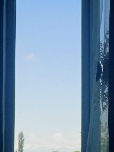 The image presents an outdoor scene viewed through what appears to be a window or a narrow horizontal opening, framed by dark bands at the top and bottom. The central and dominant feature is a wide expanse of clear, light blue sky, indicating bright daytime conditions. A faint, wispy white cloud is barely visible on the far right of the sky.
Above the central sky section, within the upper dark frame, there's a reflected or distant view of a landscape. This area shows blurred green foliage, possibly trees or bushes, with some darker, indistinct shapes that might be distant terrain or shadows. Subtle patterns resembling decorative scrolls or cursive text are visible, suggesting reflections on a textured surface or an ornate element of a window structure. On the far right of the central sky, a sliver of blurred green foliage, likely a tree, intrudes slightly into the frame, either from a reflection or a real object outside the main view.
The scene depicts a calm, bright day. No specific activity or event is taking place, and no people or identifiable objects are present beyond the natural landscape and implied architectural elements. There is no visible text. The location context is Kosh, Armenia, though the image itself does not provide specific visual cues to identify this location.