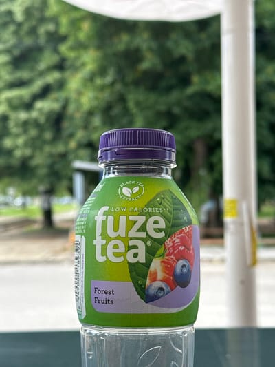 The image shows a bottle of Fuze Tea Forest Fruits. The bottle is mostly clear plastic with a green label around the middle and a purple cap. The label features images of forest fruits like strawberries and blueberries. The background is a mix of blurry green foliage and a white structure, suggesting the picture was taken outdoors on a porch or patio. The text on the label indicates it contains black tea extract, is low in calories, and urges the consumer to recycle it. No location information related to Kardam, Bulgaria is apparent in the image.