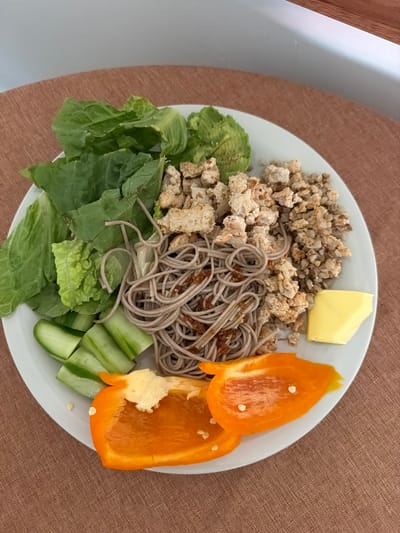 A plate of food sits on a brown surface. The plate contains buckwheat groats, a pat of butter, soba noodles, what appears to be sauteed tofu, orange bell pepper slices, cucumber spears, and lettuce leaves. A portion of a white chair can be seen on the left side of the image.
The location in Solana Beach, United States of America, cannot be ascertained solely from the content of the image. There is no contextual information to suggest where this was taken.