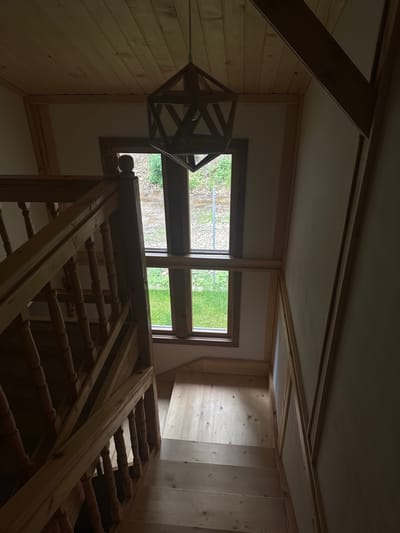 This image depicts the interior of a rustic building, likely a cabin or lodge, located in Цигов чарк, Bulgaria. The perspective looks down from an upper floor or landing, focusing on a robust wooden staircase or balustrade that dominates the foreground and midground. The balustrade is crafted from light-toned wood, featuring multiple turned spindles and sturdy banisters, suggesting a traditional or handcrafted design.
The surrounding walls are entirely clad in horizontal, light-colored wooden planks, creating a warm, natural aesthetic. Above, the ceiling is a lighter, possibly white or off-white, surface, accented by visible wooden beams and framing that segment the overhead space.
In the midground, two large windows with wooden frames offer a view of the outdoors. Through these windows, vibrant green foliage and possibly some gravel are visible, indicating a natural, possibly landscaped, exterior environment. The soft, ambient light filtering through the windows suggests it is daytime, although the interior appears somewhat dimly lit, contributing to a serene atmosphere.
A distinctive geometric light fixture, constructed from darker wood, hangs from the ceiling in the upper left part of the image. Its angular, open design adds a contemporary focal point to the traditional wooden setting. No people or specific activities are present, lending a quiet and peaceful ambiance to the scene. There is no visible text within the image. The overall impression is one of natural materials and simple, functional design, fitting for a retreat in a natural setting.