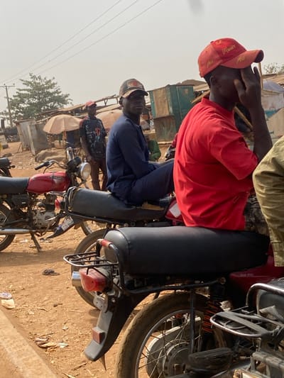 This image depicts an outdoor scene, likely a market or roadside area, characterized by unpaved, reddish-brown earth and a hazy, light-colored sky. The exact city location in Nigeria cannot be determined from the provided number "0801712591", as this is a phone number, not a geographical identifier.
In the immediate foreground on the right, the rear section of a motorcycle is visible, featuring a black padded seat, a chrome luggage rack, and a red taillight assembly. The rear wheel with spokes and a tire is partially visible.
In the mid-ground, two adult males are seated on motorcycles. The male on the right wears a red short-sleeved t-shirt and a red baseball cap with the word "MONEY" in yellow script. His right hand is raised, touching the area near his right eye. The male positioned behind him wears a long-sleeved dark blue traditional garment and a dark baseball cap. He is looking directly towards the viewer. The black seat of his motorcycle shows significant wear and tearing. To the left of these individuals, a red motorcycle with a chrome exhaust pipe, a circular headlight, and a visible engine is parked on a kickstand.
Further in the background on the left, an adult male stands upright, wearing a dark t-shirt with a red design on the chest and a red baseball cap. The environment behind the subjects consists of various rudimentary structures, appearing to be market stalls or shelters, constructed from corrugated metal sheets, wood, and tarpaulins. Several light-colored market umbrellas are also present. Overhead power lines and utility poles are visible against the sky. A deciduous tree with sparse foliage stands on the left. Scattered litter, including plastic bags and what appear to be discarded sandals, is present on the ground. No direct interactions between the individuals are discernible.
