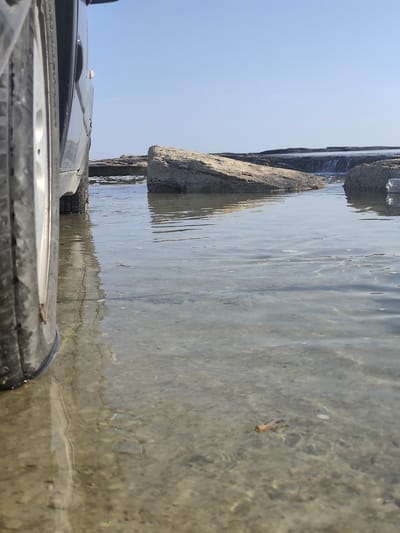 The image captures a low-angle view of the side of a dark-colored vehicle parked in shallow water. The vehicle's tire is prominently visible on the left side of the frame, partially submerged. In the background, there are large, light-colored rocks scattered in the water, and a small waterfall or cascade is visible over a rocky ledge. The sky above is clear and blue. The environment appears to be a natural body of water, possibly a shallow sea or a large river with rocky outcrops, near the city of Saiyn, Kazakhstan. There are no people present in the image. The water is clear enough to see the sandy or rocky bottom in some areas, and there are some ripples and small bubbles on the surface.