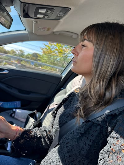 A woman with light brown hair is seated in the driver's seat of a vehicle. She is wearing a black lace top and a seatbelt. Her head is tilted downwards, looking at something out of the frame. Outside the car window, trees and a fence are visible. The location is Saint-Laurent-du-Var, France.