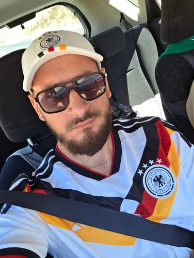 A man is shown in a selfie taken from a slightly high angle, with the camera positioned to capture his chest and face. He is wearing a white jersey with the German flag colors (black, red, and yellow) as stripes across the chest, and a circular emblem on the left side. The jersey also features black stripes on the sleeves. He is also wearing a white baseball cap with a similar emblem and colorful squares on the brim, which appear to be the German flag colors. Dark-rimmed sunglasses cover his eyes. He has a beard and is wearing a thin gold chain. The man is seated in a vehicle, evidenced by the seatbelt across his chest and the car seats visible behind him. The background suggests a bright, outdoor setting, possibly a sunny day, with a hint of arid landscape visible through the car window on the right. The image appears to be taken in a location with a warm climate. The overall mood conveyed is casual and possibly celebratory, given the sports-themed jersey. No specific text is clearly legible beyond the emblem on the jersey and cap. The image appears to have been taken during daylight hours. The context provided indicates the location is Ramat Negev Regional Council, Israel.