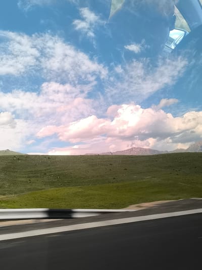 The image presents a scenic landscape captured from the perspective of a moving vehicle in Yelpin, Armenia. The upper two-thirds of the frame are dominated by a vast blue sky adorned with an array of white and pink-hued clouds, ranging from wispy cirrus to large, voluminous cumulus formations. The soft pink tones on some clouds suggest a particular atmospheric condition or possibly the time of day, though the bright overall illumination indicates daylight.
Below the sky, a wide expanse of rolling green hills stretches across the middle ground. These hills are covered in lush, low-lying vegetation, appearing largely treeless and undulating. In the far distance, rugged mountains are visible, some exhibiting lighter patches that suggest snow caps or exposed rock formations, adding to the grandeur of the landscape.
The immediate foreground features elements indicative of travel: a dark asphalt road with a distinct white lane marking on the right, and a white metal guardrail on the left. Both the road and guardrail show signs of motion blur, confirming that the photograph was taken from a vehicle in transit. Reflections are discernible in the upper right corner, likely from a car's windshield or window, including a partial view of a side mirror and a faint outline that could be a person's head or shoulder.
The scene conveys a sense of open-road travel through a beautiful, natural environment, characterized by expansive skies, verdant hills, and distant mountains. The weather appears clear and bright. There is no visible text in the image.