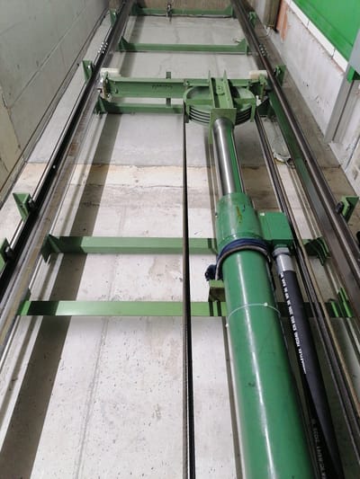 This is a forensic, matter-of-fact description of the provided image, which appears to be an elevator shaft in Grünwald, Germany.
**Scene Description:**
The image captures a detailed view of an elevator shaft. The shaft is lined with industrial green metal beams and supports, forming a framework for the elevator's operation. Two parallel steel rails, one on each side of the shaft, are visible, serving as guides for the elevator car. Multiple steel cables, thick and twisted, hang vertically within the shaft, presumably for hoisting and safety mechanisms.
Dominating the foreground and extending into the mid-ground is a large, cylindrical green hydraulic actuator. This actuator is positioned vertically within the shaft and is connected to what appears to be a pulley system and other mechanical components mounted on the green beams. A thick black hose, with white lettering that includes "HANSALFLEX HD240 EN 853 2SN WP 90 BAR", is attached to the actuator, indicating it's part of the hydraulic system. The hose is secured to the actuator with a blue strap and white zip ties.
The walls of the shaft are constructed of rough, grey concrete. The floor of the shaft is also concrete, showing signs of wear and tear, with some debris and staining. Shadows are cast across the floor and some of the metal structures, suggesting an overhead light source.
The text "Grünwald" is visible, handwritten in black marker on one of the green metal beams in the upper left portion of the image, confirming the location. No people are visible in the image. The overall environment is industrial and functional, characteristic of an operational elevator shaft.