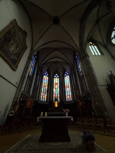The image depicts the interior of a church, likely the Saint-Louis Church in Ribeauvillé, France, given its architectural style. The perspective is wide-angle, looking towards the altar area. The vaulted ceiling with Gothic architectural elements is prominent in the upper portion of the image, with dark stone arches. On the left wall, a large, ornate framed painting hangs. The central focus is on the altar, which is draped with a white cloth and adorned with two lit candles and a vase of pink flowers. Behind the altar, a large stained-glass window, illuminated by natural light, displays vibrant colors and intricate patterns. Additional stained-glass windows are visible on the side walls, casting colorful light onto the stone pillars. In the foreground, a patterned rug covers the wooden floor, and rows of wooden chairs are arranged on either side of the aisle. To the right of the altar, a wooden crucifix is mounted. The lighting is dim, with the primary illumination coming from the stained-glass windows. There are no people visible in the image.