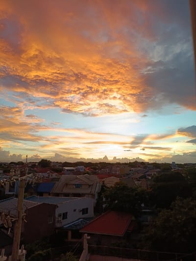 The image shows a view of a residential area in Parañaque, Philippines, taken from a rooftop. The sky is a vibrant mix of orange, yellow, and blue hues, with fluffy clouds creating a dramatic sunset effect. The sun, positioned behind the clouds, casts a warm glow over the cityscape. The rooftops of houses and apartment buildings are visible in the foreground, interspersed with patches of green trees. A utility pole with wires stands tall amidst the buildings, adding a sense of urban life to the scene. The scene is captured from a perspective slightly above the roof level, allowing for a clear view of the sky and the surrounding buildings.  The coordinates provided are: 14.474, 121.008.