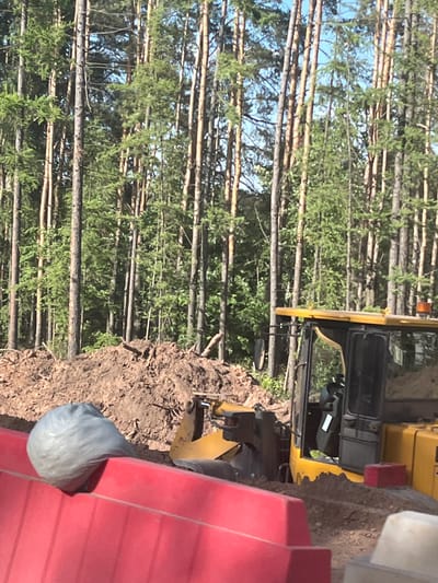 The image shows a yellow construction vehicle parked next to a pile of dirt and a forest of tall, thin trees. A grey plastic bag is visible near the dirt pile, adjacent to a red object with horizontal ridges. The location is likely Staryi Chul'tem, Russia.