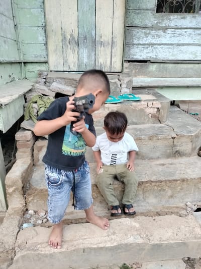 The image depicts two young male children on rough concrete steps outside a weathered wooden house in Cutneuheun, Indonesia. The child on the left, standing barefoot on a lower step, wears a black t-shirt with a "SURABAYA" graphic and faded denim shorts. He holds a black toy gun with both hands, pointing it forward and slightly downward, his gaze following the toy's direction. The child on the right sits on a higher, cracked concrete step, wearing a white t-shirt with a "PUMA" graphic, olive-green pants, and black sandals. He is looking down, his hands resting on his knees. In the background, an aged wooden house features vertical planks painted faded light green and teal, with significant paint peeling. A closed wooden door is centrally located, and an upper window with a metal grate is visible on the right. Two teal flip-flops rest on an upper concrete step to the right. A crumpled light green cloth lies on the ground to the far left. The concrete steps exhibit deterioration and exposed brickwork.
