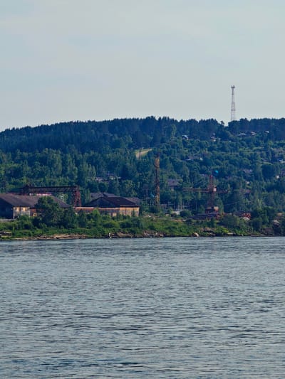 The image shows a wide river in the foreground, with a densely forested hill rising steeply in the background. Along the base of the hill, a line of industrial buildings and infrastructure is visible, including warehouses, cranes, and a tall communications tower. Scattered among the trees on the hillside are houses, indicating a settlement. The sky above is clear and pale blue. This scene is characteristic of the city of Predivinsk, Russia, a settlement located on the Yenisei River.