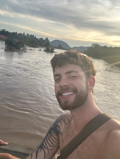 The image presents a selfie featuring a single male subject in the foreground against a natural backdrop. The man has curly brown hair and a beard, is smiling with visible teeth, and is shirtless. He wears a silver ring piercing in his right nostril, a silver barbell piercing above his left eyebrow, and a silver hoop earring in his left earlobe. A large, dark tattoo with intricate patterns covers his left shoulder and upper arm. A dark olive-green strap, possibly from a bag, is visible across his right shoulder.

The background consists of a wide body of brownish-tan water, indicating a river, with visible ripples and current. On the distant left bank, structures with red roofs are discernable among dense green foliage, including palm trees, consistent with a settlement in Khorn Neua, Laos. Further in the distance, a range of dark green mountains or hills is visible under a sky characterized by scattered light clouds. The scene is illuminated by warm, low-angle sunlight, resulting in a prominent lens flare in the upper right quadrant of the image.