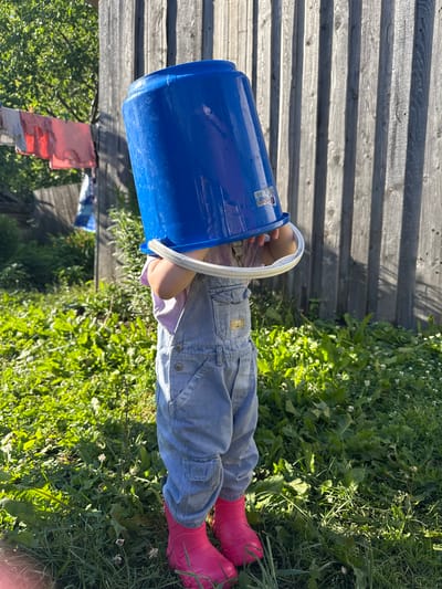 A full-body shot captures a toddler in a backyard in Varzi-Omga, Russia, with a blue bucket on their head. The child is wearing light blue denim overalls over a pink shirt, and bright pink rubber boots. The child is standing in a grassy area with lush green foliage. Behind the child, a weathered wooden fence runs vertically. Clothes hang on a clothesline to the left of the frame, with trees and other greenery visible in the background. The lighting suggests it is daytime, with sunlight casting shadows.