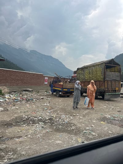 Two women stand in a dirt lot covered in litter. One woman, on the left, wears a gray headscarf and matching outfit. The other woman, on the right, wears an orange headscarf and a peach-colored outfit. Both women are holding their hands to their faces. Behind them are two trucks; one is blue and yellow, and the other is a large, brown truck with a yellow tarp covering its cargo. A brick wall runs along the left side of the image, and in the background, mountains rise under a cloudy sky. The scene appears to be in a rural or semi-rural area, possibly a roadside dumping ground. The litter includes plastic bags, paper, and other debris.