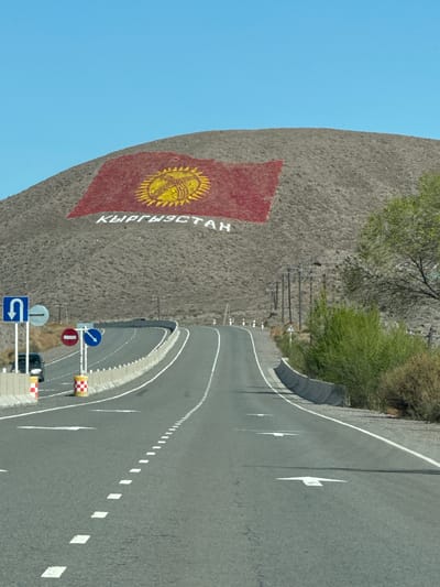 The image depicts a road leading towards a hillside in Kok-Moinok - 1, Kyrgyzstan. The hillside is painted with a large representation of the Kyrgyzstan flag and the country's name. Roadside barriers and signage are visible along the asphalt road. The sky is clear and blue.