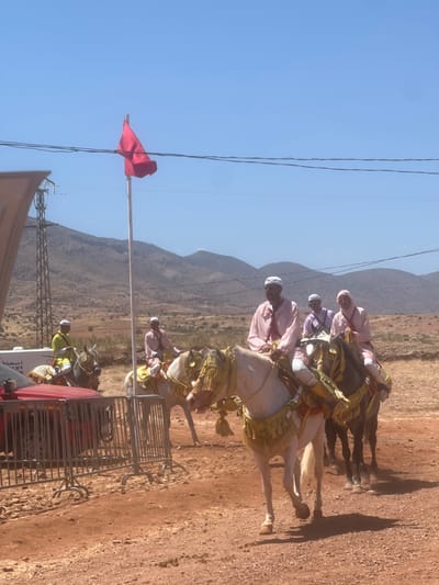 This image depicts a procession of people on horseback in Tabia, Morocco. The scene is set against a backdrop of arid hills and a clear blue sky. A red flag is prominently displayed on a flagpole. Several riders, adorned in traditional pink and white attire, are mounted on horses. The horses themselves are decorated with ornate, golden saddle blankets and tassels. In the foreground, a red car is visible behind a metal barrier. The ground is covered in reddish-brown soil. The overall impression is of a cultural or ceremonial event taking place in a rural Moroccan setting.
