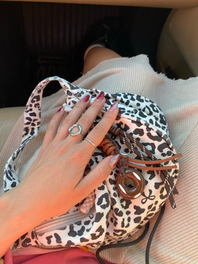 A person's hand is resting on a leopard-print bag. The hand has two rings on the fingers: one silver ring with an irregular shape and another thin, textured ring. The fingernails are long and painted with a design that transitions from pink to red. The bag has brown leather detailing and a donut-shaped decorative element. The bag sits on a light-colored fabric, possibly clothing. A small portion of what appears to be a black shoe is visible on the left side. The setting appears to be inside a vehicle. There is no indication of the image being set in El Bruc, Spain.