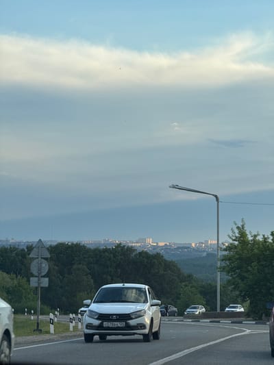The image depicts a cloudy sky with a faint sunset glow, casting a soft light over a landscape. A city skyline is visible in the distance, nestled amongst rolling hills covered in dense green trees. The foreground is dominated by a road, suggesting an outdoor setting, possibly a highway or a scenic route. Several cars are present, including a white car with visible Russian license plates. The license plate reads "G578BA 126". A street lamp on a pole is also visible, indicating urban or suburban proximity. The overall mood is tranquil, with the diffused light and distant cityscape creating a sense of calm. The time of day appears to be late afternoon or early evening, judging by the lighting conditions and the faint sunset hues in the sky. The weather is predominantly cloudy, but there are breaks in the clouds allowing some light to filter through. The location is identified as Novomar'evskaya, Russia.