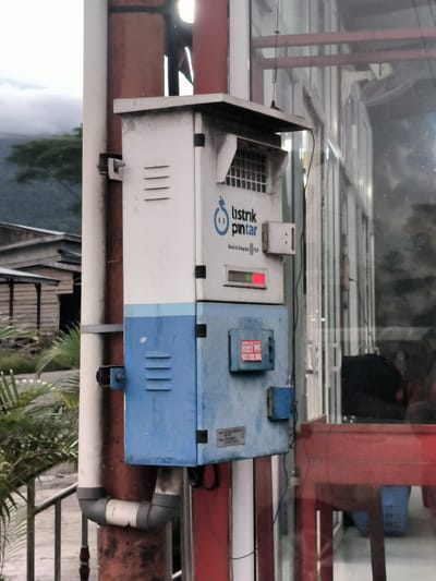 The image displays an electrical meter box, likely a prepaid "smart electricity" unit, mounted outdoors on a pole. The box is bipartite, with an upper white section and a lower light blue section, featuring ventilation slats and a small antenna on top.

Prominently displayed on the white section are the words "listrik pintar" (smart electricity) and "Solusi Isi Ulang dari PLN" (Recharge Solution from PLN), identifying it as part of Indonesia's state-owned electricity utility system. A small red indicator light is visible on the white panel. On the lower blue section, a red sticker advertises "KARISMA JAYA SEDOT WC SUMUR BARA" (septic tank/well emptying service) along with a phone number (0822 7756 7885).

The electrical box is secured to a reddish-brown vertical pole, which also supports white PVC piping with grey elbow joints near the bottom. To the right of the pole, a large glass window reflects a red table or counter and what appears to be a blue basket from the interior of a building.

The background on the left side of the frame features rustic wooden structures, green foliage resembling palm fronds, and distant mountains partially veiled by clouds or mist, suggesting an overcast day. No individuals or specific activities are visible in the scene. The setting blends modern utility infrastructure with a semi-rural landscape in Sukamulia, Indonesia.