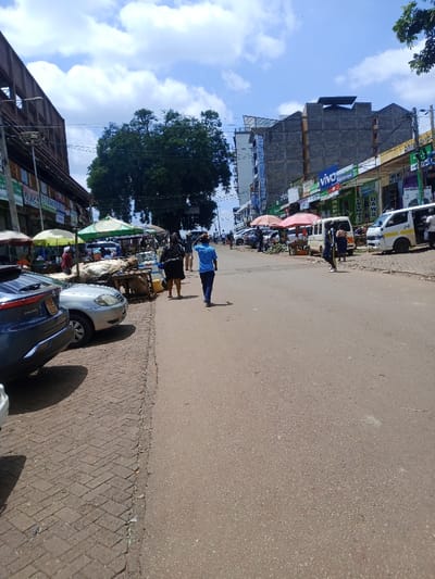 The image displays a street scene in Murang'a, Kenya, under a partly cloudy blue sky. The main element is an asphalt road stretching into the distance.

On the left side of the road, a brick-paved sidewalk is present. A dark blue SUV is parked with its rear towards the viewer, partially obscuring a silver sedan parked behind it. Along this sidewalk, several market stalls operate under colored umbrellas (green, yellow, red), featuring tables displaying goods. Behind these stalls, multi-story buildings with numerous shopfronts are visible. A large, dense green tree stands in the mid-ground, left-center.

On the right side of the road, a continuous line of multi-story commercial buildings is visible. Some buildings feature exposed concrete and glass elements. Prominent commercial signs, including a "VIVO" logo, are displayed. Below these buildings, additional market stalls with umbrellas are set up. Several white vans, consistent with matatus, are parked or in motion along this side of the street.

In the foreground and mid-ground of the road, two individuals are walking away from the camera: one person in dark attire on the left, and another person wearing a blue shirt and dark pants in the center. Additional people are visible throughout the scene, either walking, standing, or engaged with the market stalls and vehicles on both sides of the street. Overhead, power lines and utility poles are present. The scene is illuminated by daylight.
