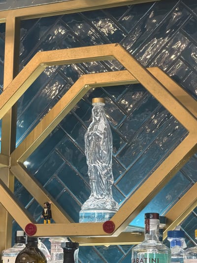 The image is a close-up shot of a liquor cabinet located in Tarquinia, Italy. The cabinet appears to be made of gold-colored metal framing around a backdrop of blue, glossy, rectangular tiles laid in a herringbone pattern. The cabinet shelves hold several liquor bottles, including a long, clear bottle with a gold-colored cap, and a bottle of Sabatini gin. A miniature figure dressed in black and yellow is visible on one of the shelves.