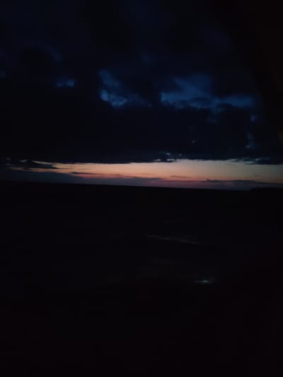 The image depicts a dark, atmospheric landscape, likely taken at dusk or night. The sky is dominated by heavy, dark clouds, with hints of twilight colors—pinks and oranges—peeking through near the horizon. The lower half of the image is almost entirely black, suggesting a ground or water surface devoid of significant light sources. There are faint, greenish-blue lights visible in the dark area, possibly distant artificial lights or a reflection. There are no discernible people, objects, or actions in the scene. The environment appears to be rural or undeveloped.