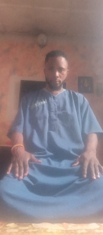 A man is seated with legs crossed, wearing a blue shirt with small buttons at the top. His hands rest on his lap. A gold bracelet adorns his left wrist. Behind him is a wall with a framed picture hanging slightly to the right. A television is also visible behind him. An overhead light fixture is seen near the top of the frame.