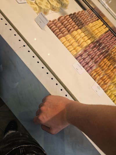 A person's arm and hand are visible in the foreground of the image, positioned over a white display counter. The counter showcases rows of colorful macarons, with labels indicating flavors such as "MACARON CASSIS" and "MACARON CARAMEL," each priced at "1650". In the background, out of focus, are what appear to be pastries on the same counter. The environment suggests a retail setting, possibly a bakery or patisserie, located in Chevilly-Larue, France. The lighting is bright, casting shadows on the display and the person's arm.