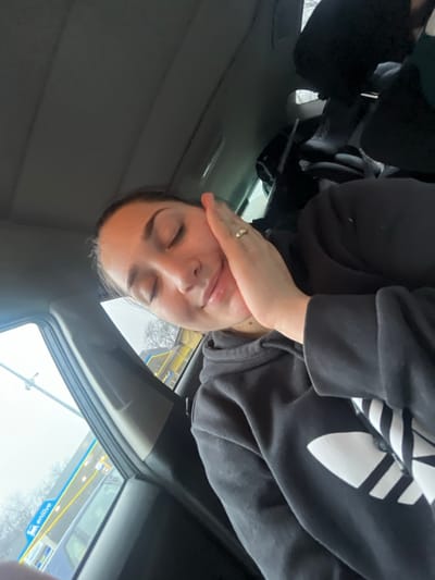 The image depicts a female individual resting inside a vehicle, parked at an Enilive fuel station in Lonate Pozzolo, Italy. The subject, positioned centrally and angled, has dark hair, closed eyes, and a slight smile, with her right hand gently placed against her right cheek. A ring is visible on her right ring finger. She is wearing a black hooded sweatshirt featuring a large white Adidas trefoil logo on the front. The vehicle's grey headliner and dark interior components are visible. Through the car's side window, the distinctive blue and yellow canopy of the Enilive station, marked with the "enilive" logo, is clearly visible under an overcast sky. A bare tree and a partial view of another vehicle are present in the exterior background.