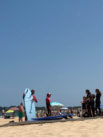 On a sunny day at a sandy beach in Ribamontán al Mar, Spain, a surf lesson or instructional session is in progress. In the midground, a male individual wearing a red long-sleeved wetsuit and sunglasses holds a light blue surfboard vertically. To his right, another person in a red wetsuit and a red cap stands. Further right, a group of approximately seven individuals, primarily children, all wearing dark full-body wetsuits, are gathered attentively. Two dark blue surfboards lie horizontally on the sand in front of this group, resting on a wooden mat. To the left of the central figure, a shirtless man in green swim trunks stands. The background features numerous other beachgoers distributed across the sand, some under colorful beach umbrellas. The upper portion of the image is dominated by a clear, light blue sky. Distant coastal vegetation is visible on the horizon.