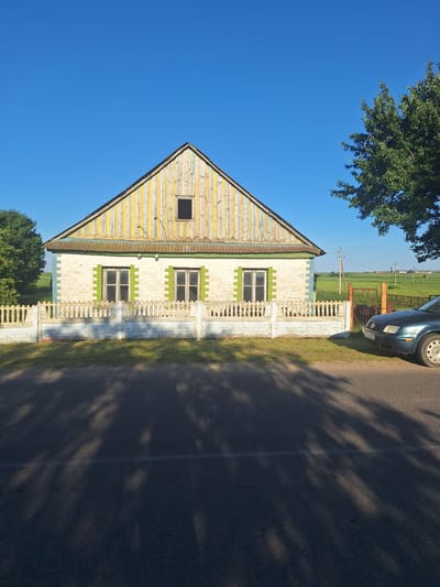 The image shows a single-story house with a gabled roof, located in Цімкавіцкі сельскі Савет, Belarus. The house has a white brick exterior with green accents around the windows. The roof is made of weathered, vertical wooden planks painted in alternating yellow and white stripes. A small, dark square window is visible in the attic. A low, decorative fence made of white brick and posts surrounds the front of the property. Two windows with green shutters are visible on the front of the house. A dark-colored car is parked on the right side of the image, partially obscuring a metal gate and fence leading to a green field. Trees are present on the left and right sides of the house, and utility poles are visible in the background. The sky is clear and blue, and shadows from trees are cast across the asphalt road in the foreground.
