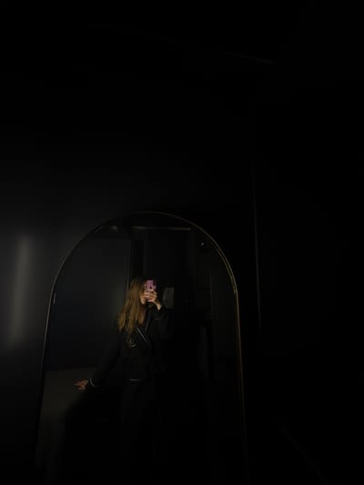 The image shows a reflection in a round mirror within a dark, possibly indoor, setting. The reflection reveals a person holding a pink mobile phone in front of their face. The person has long, blonde hair and is wearing a dark-colored outfit with white trim. The environment is largely undefined due to the darkness, but the mirror's surroundings appear to be dark walls. The city of Ciudad López Mateos, Mexico is mentioned in the prompt, but there is no information in the image to confirm that this is where the image was taken.