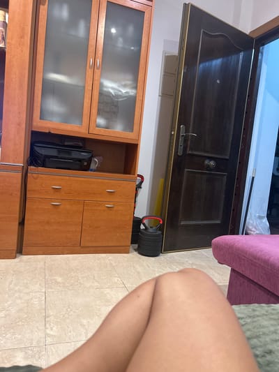 The image depicts an interior scene, likely a living room or common area, in a residence in Pueblo Salinas, Spain. A wooden cabinet unit with frosted glass doors is prominently featured on the left. The cabinet contains a black printer on its top shelf. Below the upper cabinets are drawers and two cabinet doors, all with silver handles. To the right of the cabinet, a dark wooden door is ajar, revealing a glimpse of a brightly lit space beyond. Next to the door, a stack of black weight plates with red handles and a black mop with a white cloth at the end are visible. In the foreground, a portion of a person's legs and knees are visible, positioned on a tiled floor. A portion of a magenta-colored ottoman or sofa is also in the lower right of the frame. The environment appears to be indoors, with tiled flooring and neutral-colored walls.