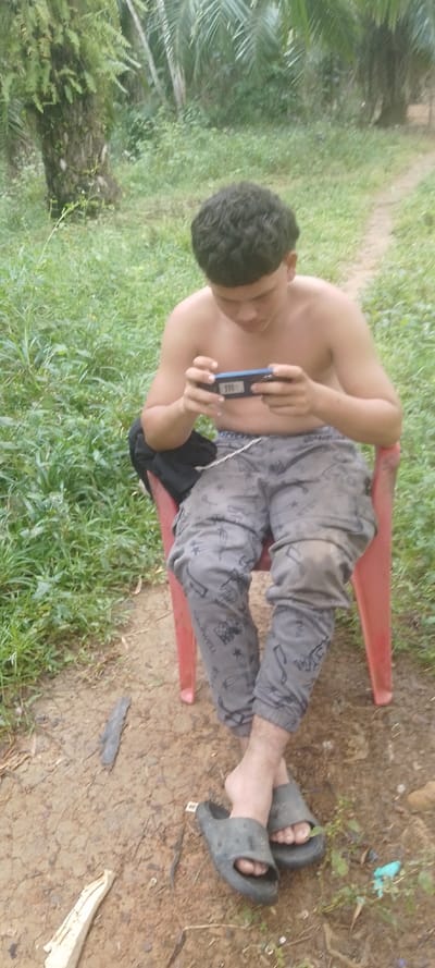 A shirtless young man with curly hair sits in a red plastic chair in a grassy outdoor setting. He is engrossed in a mobile phone game, holding the device horizontally. He wears patterned gray pants and dark-colored sandals. The ground is a mix of dirt and vegetation. In the background, there are trees and greenery suggesting a rural or natural environment.
