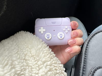 A close-up image captures a person's right hand holding a lavender-colored silicone case designed to resemble a retro video game controller. The case features a white directional pad on the left, two white circular buttons with cat paw prints, and four white action buttons labeled 'Y', 'X', 'A', 'B' on the right, along with a small central indentation. Small, rounded protrusions resembling ears are visible on the top edge of the case. The person's fingernails are manicured to a short-to-medium length and covered in a light pink, glittery polish. A section of white, textured, fluffy fabric is prominent in the lower left foreground. The background consists of dark, indistinct fabric in the upper left, while a dark, structured object with visible stitching, consistent with a bag or vehicle interior component, appears on the right. Light blue fabric, likely denim, is visible beneath the hand. The scene is located in the city of Липицы, Russia.