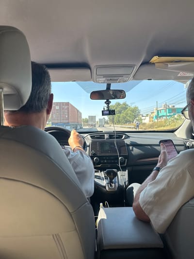 From inside a car, a seated passenger rests their head against the headrest of the front seat. A driver holds the steering wheel. Another seated person in the passenger seat looks at their phone. The vehicle is in transit, evidenced by the view through the windshield: urban scenery with buildings, overhead power lines, and pedestrians on the sidewalk. A dashcam is mounted under the rearview mirror. The car’s interior is light gray.