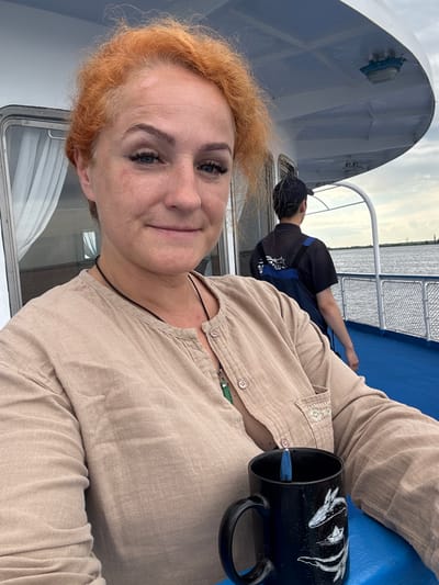 A woman with ginger hair is taking a selfie on the deck of a boat. She is wearing a beige shirt with buttons and a necklace. She is holding a black mug with a blue spoon in it. In the background, another person wearing a black cap and shirt is standing near the railing, looking out at the water. The boat appears to be on a river or a lake, with a cloudy sky overhead. The location is the city of Menzelinsk, Russia.
