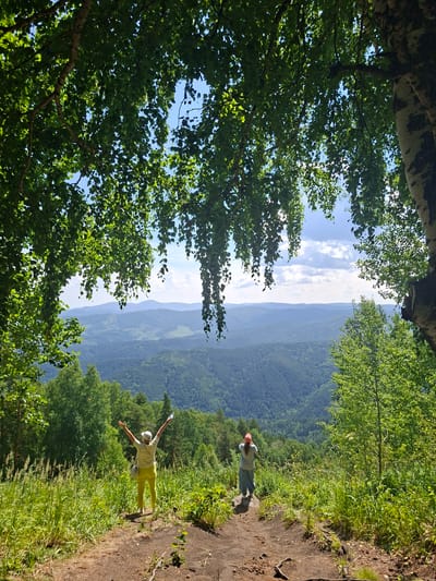 The image depicts a vast, natural landscape in Belokurikha, Russia. In the foreground, lush green trees with dense foliage frame the scene, with their branches and leaves creating a natural canopy. Sunlight filters through the leaves, suggesting a clear, bright day. Beyond the trees, a steep, verdant mountain range stretches into the distance, its slopes covered in a thick forest. The layers of mountains fade into a hazy blue in the far distance, indicating a significant depth to the vista.

Two individuals are visible on a dirt path that winds along the edge of the mountain. The person in the foreground, wearing a light-colored shirt and bright yellow pants, has their arms outstretched, seemingly embracing the expansive view. They appear to be in a relaxed and joyful pose. Further up the path, another individual in a light-colored shirt and a red hat stands with their hands behind their back, also facing the landscape. The path itself is rough and earthy, with scattered stones and patches of grass growing along its edges, suggesting a hiking or walking trail.

The overall impression is one of serene natural beauty and outdoor recreation. The time of day appears to be daytime, with bright sunlight casting shadows, and the weather seems fair and pleasant. The location, Belokurikha, Russia, implies a mountainous resort area known for its natural scenery. There is no visible text in the image.