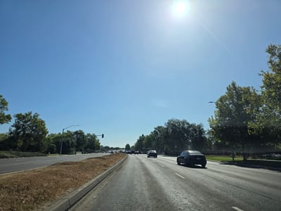 This is a daytime, outdoor photograph taken from a vehicle's perspective, likely on a sunny day given the bright sun in the upper right corner of the image and the clear blue sky. The scene is a multi-lane road with traffic. Trees line both sides of the road, with lush green foliage indicating a warm season. On the right side of the road, there is a dark-colored sedan. On the left side, there is a grassy median with some dry, brown vegetation along the edge of the road. Traffic lights are visible in the distance, with at least one showing a green light. The road appears to be in an urban or suburban area, possibly in Elk Grove, United States, given the location context. The overall impression is of a typical day driving on a highway or a major road. There is no visible text in the image.