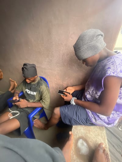 Two individuals are seen from above, occupied with their phones. One wears a gray beanie, a purple top, and navy shorts, while the other sits in a blue plastic chair, sporting a black head covering, an army green t-shirt, and black shorts. Both are intensely focused on their devices. Additional bare feet and lower body parts of other people are partially visible around them, suggesting a crowded space.